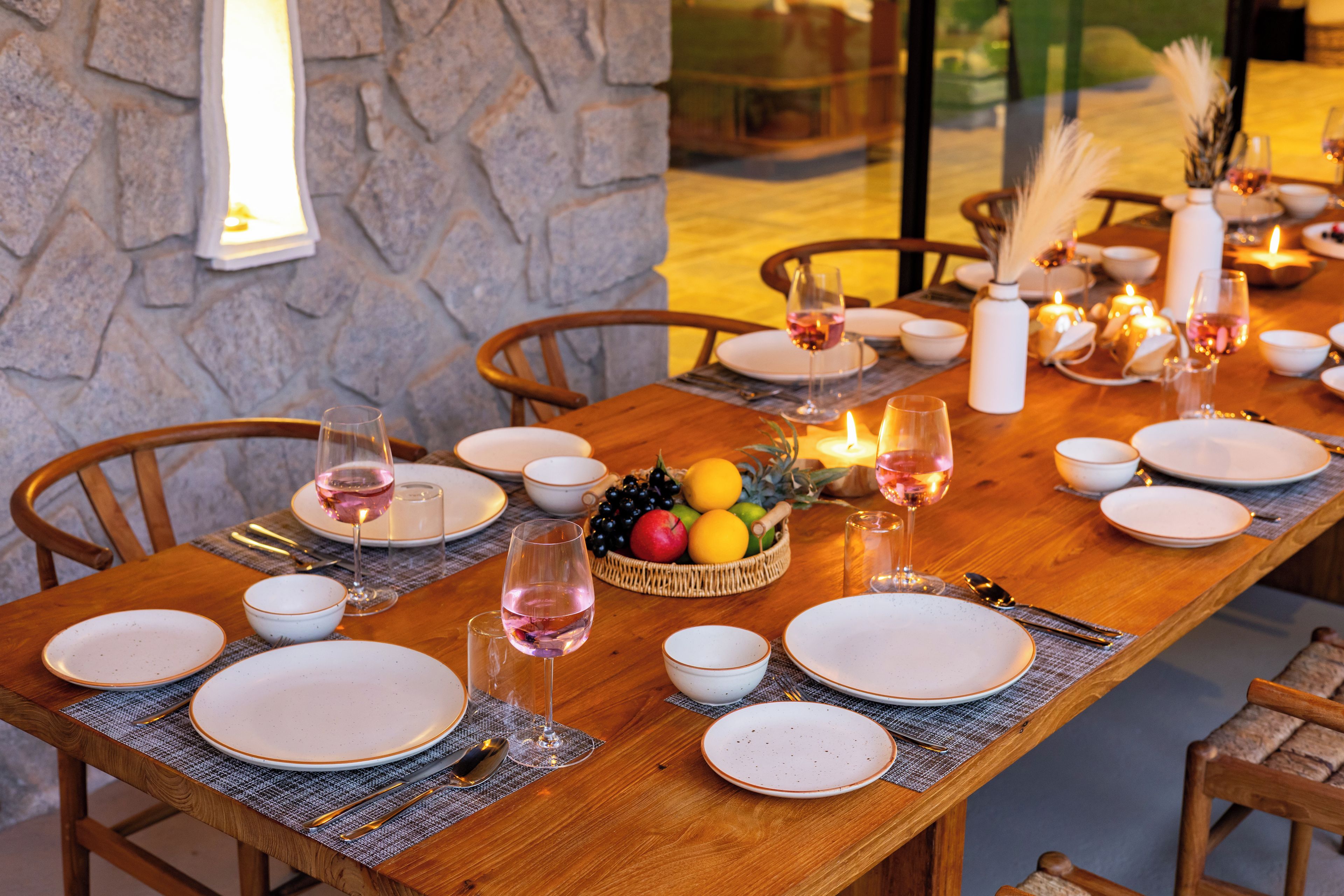 Una mesa de comedor dispuesta con platos, vasos y una cesta de frutas, rodeada de sillas.