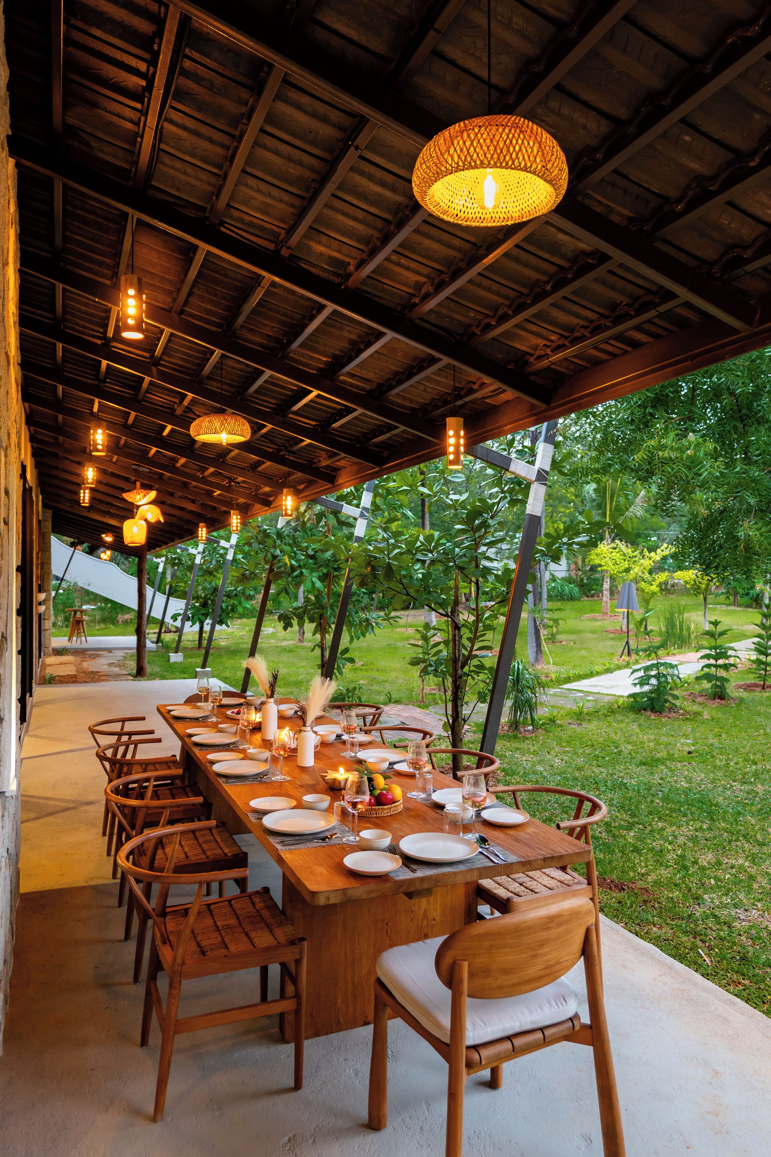 An outdoor dining area with a long wooden table set for a meal, surrounded by chairs, under a wooden ceiling with hanging lights.