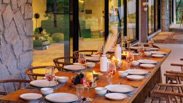 Una larga mesa de comedor preparada para una comida, con sillas a ambos lados, ventanas amplias y lámparas colgantes.