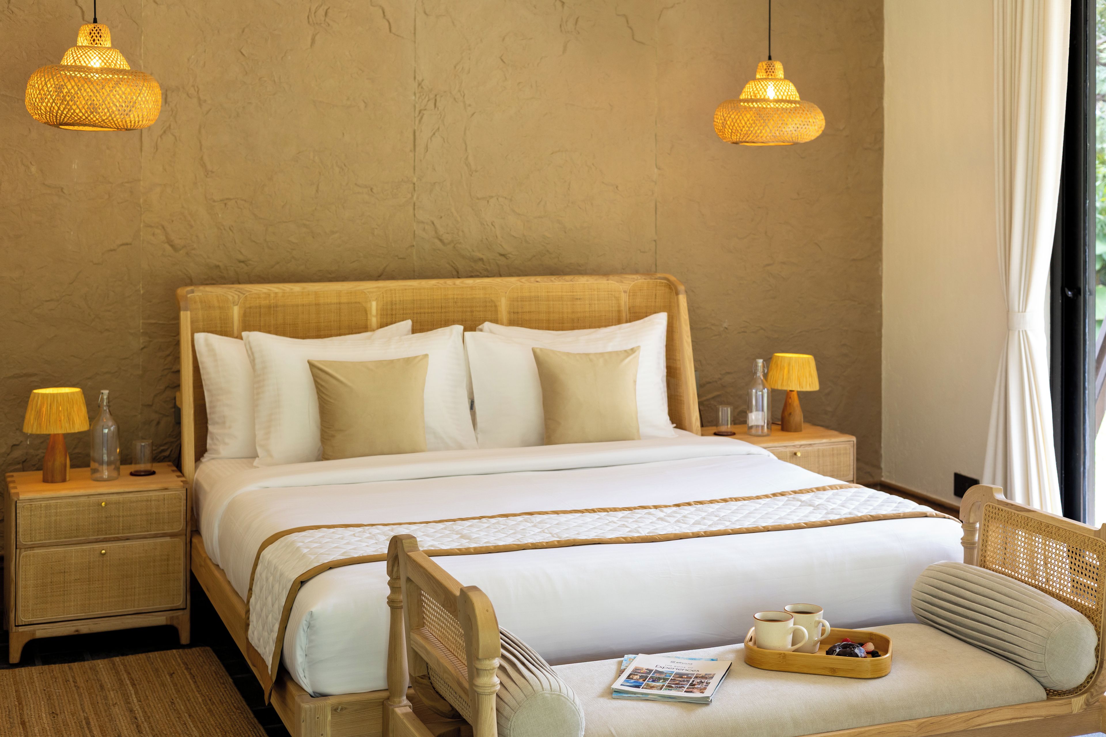 Habitación de hotel con cama de madera, dos lámparas de noche, una mesita de noche, una silla de mimbre, una bandeja con tazas y una revista, y una ventana con cortinas.