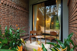 A cozy outdoor seating area with a brick wall, two wooden chairs, a small table, and a glass door leading to a bedroom with a bed and a nightstand. - ELIVAAS Prive Saukhyam 3BHK Villa (Bengaluru)