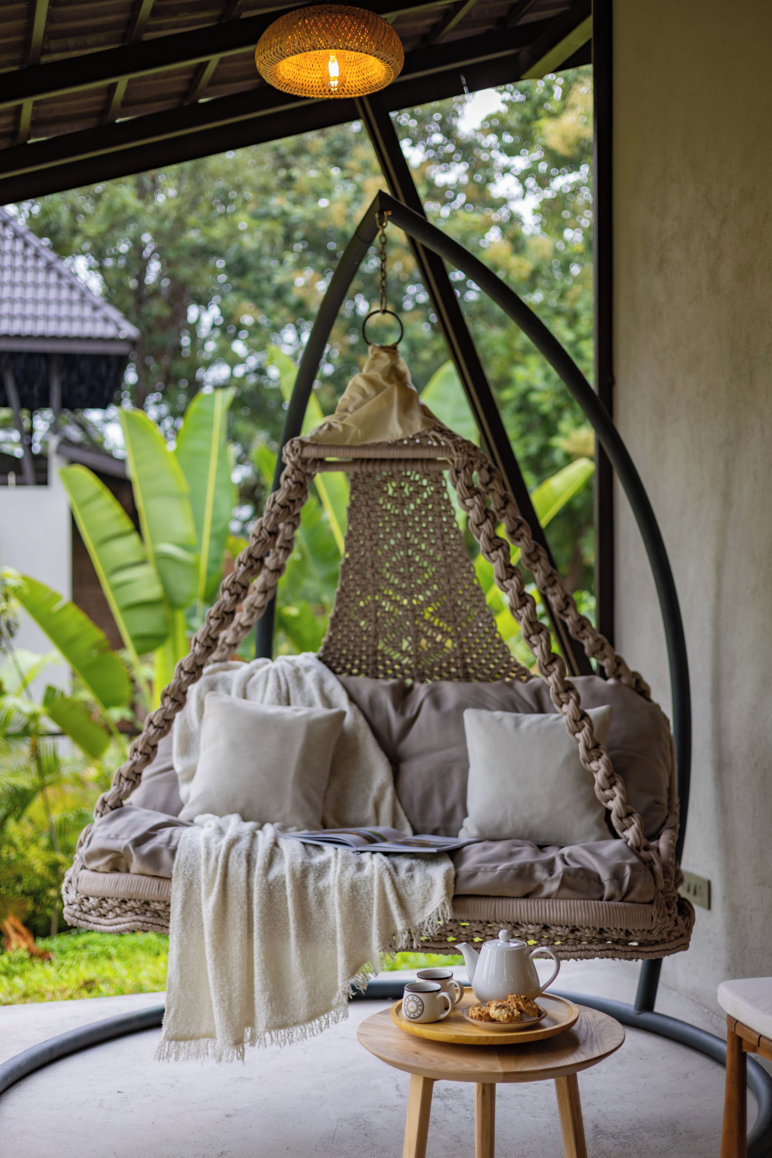 A hanging swing chair with cushions and a throw blanket, a small round table with a teapot and cups, and a view of greenery outside.
