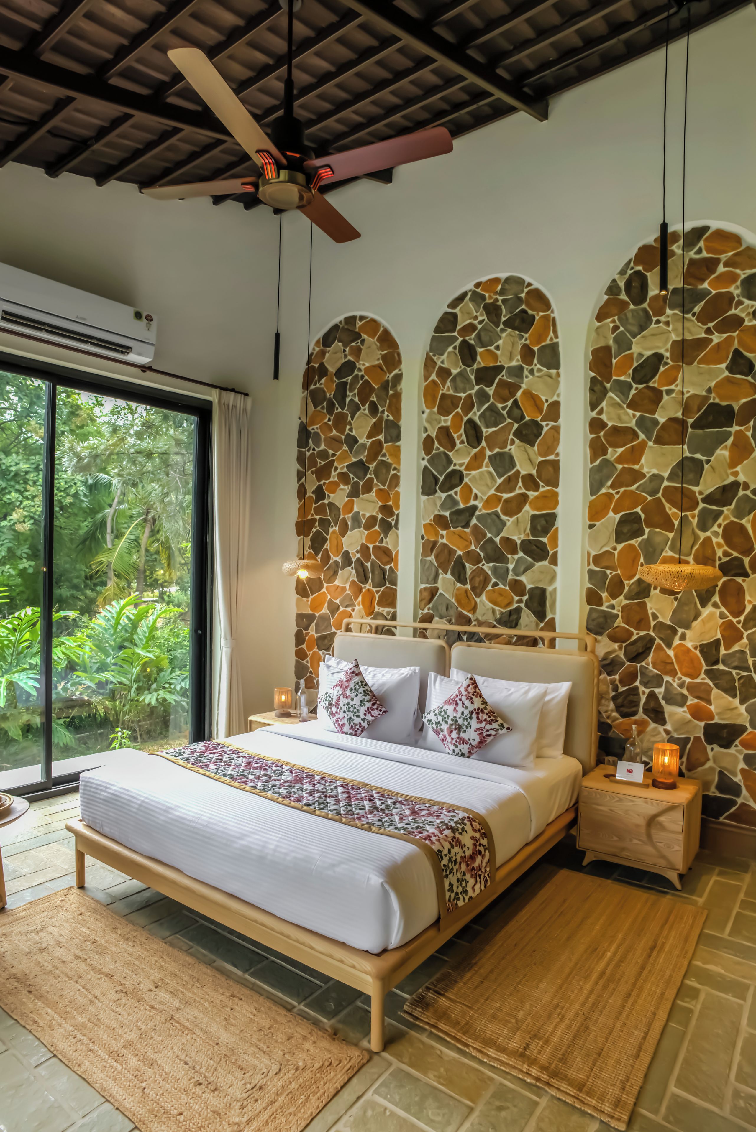 A bedroom with a bed, a nightstand, a ceiling fan, and a stone wall.