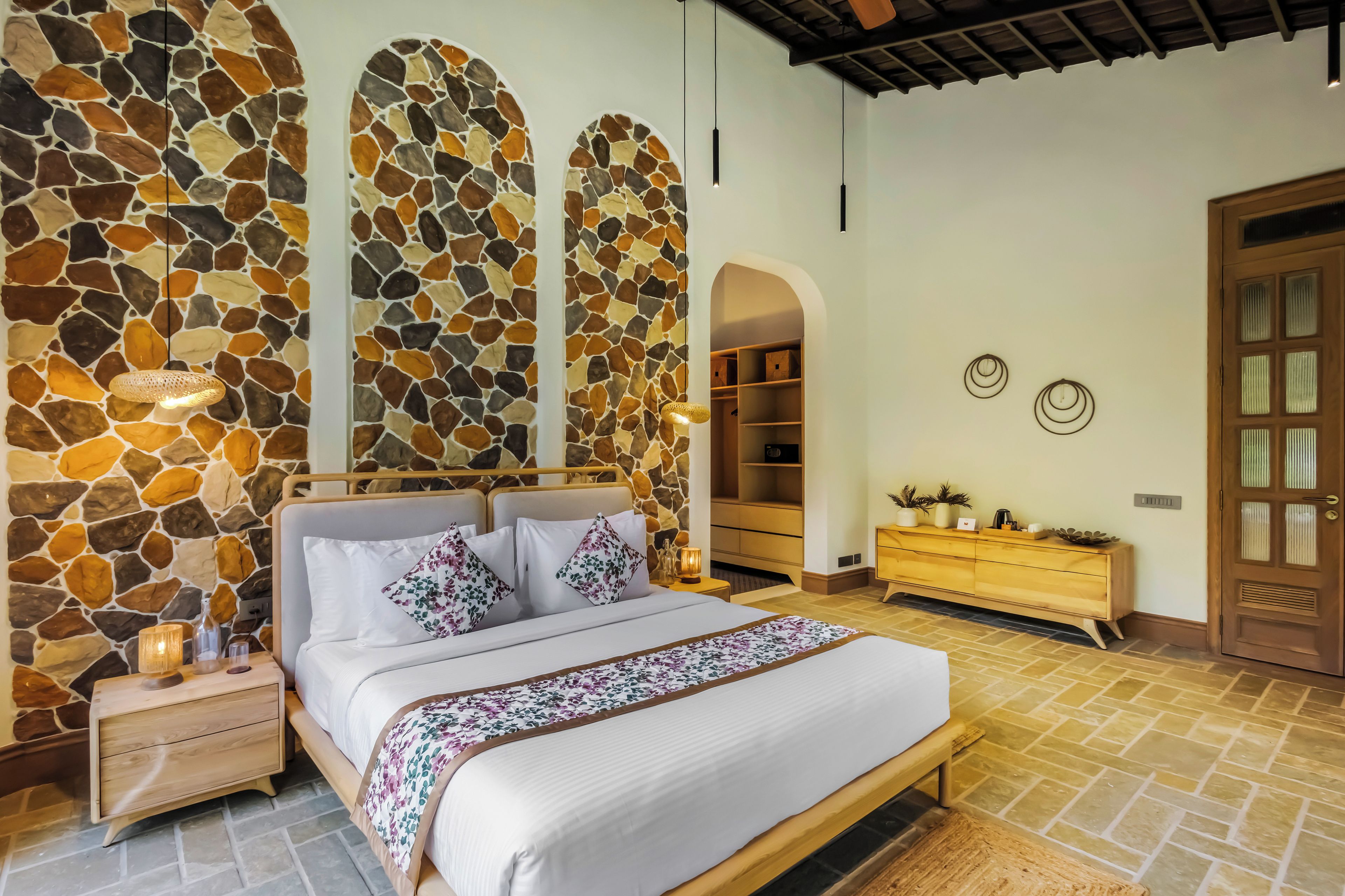 A bedroom with a stone wall, a bed with floral pillows, a wooden dresser, and a door with glass panels.