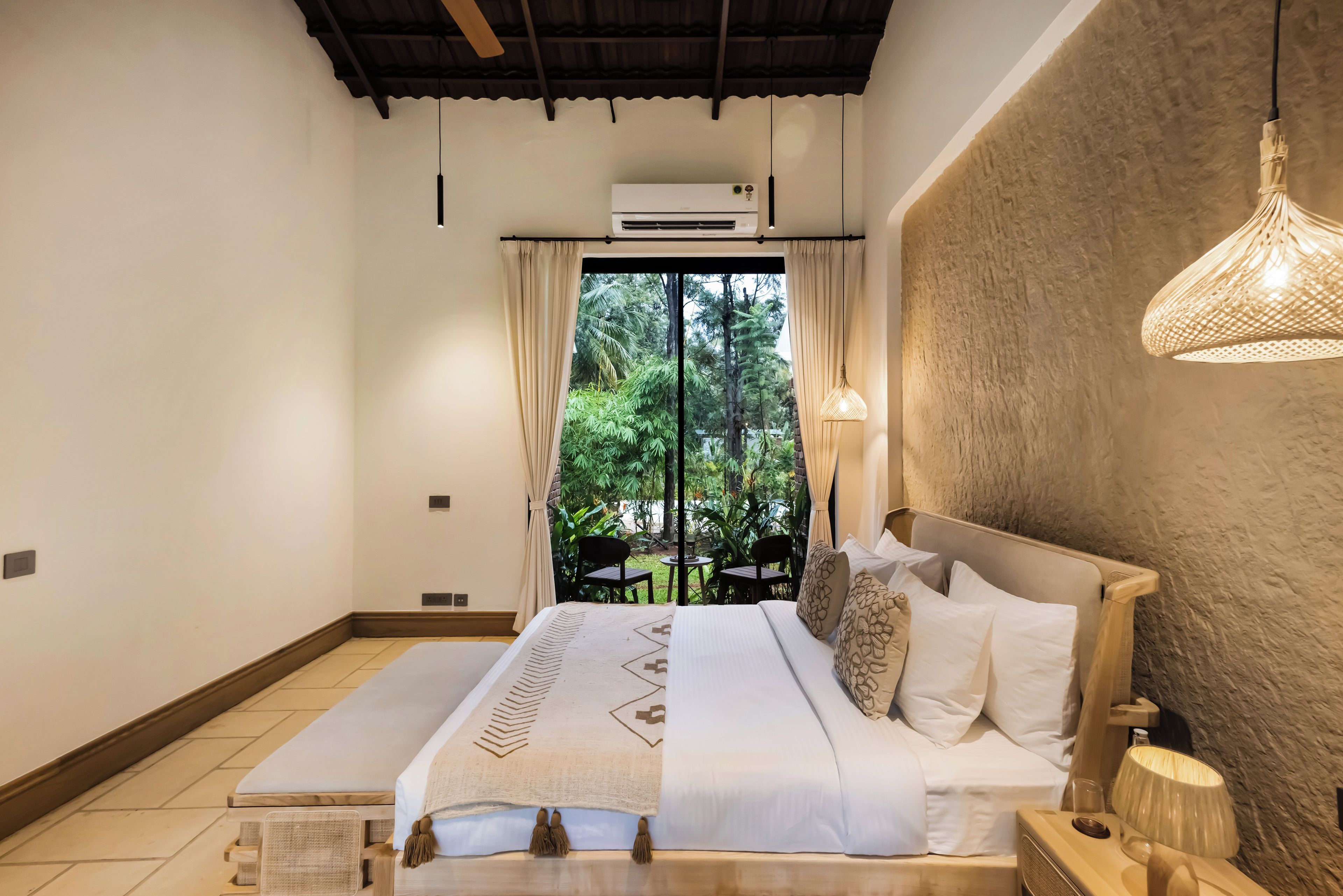 A bedroom with a bed, a nightstand, a lamp, an air conditioning unit, and a view of greenery outside.