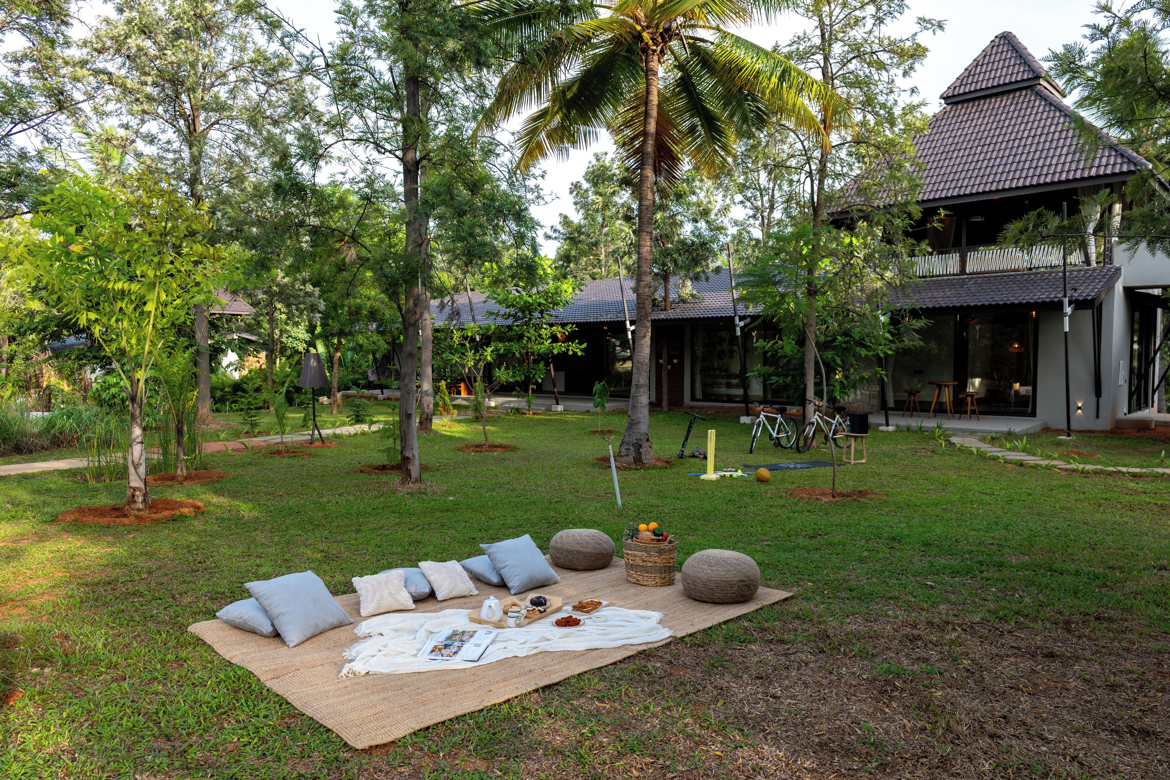 A serene outdoor setting with a picnic setup on a grassy area, surrounded by trees and a modern building with a tiled roof.