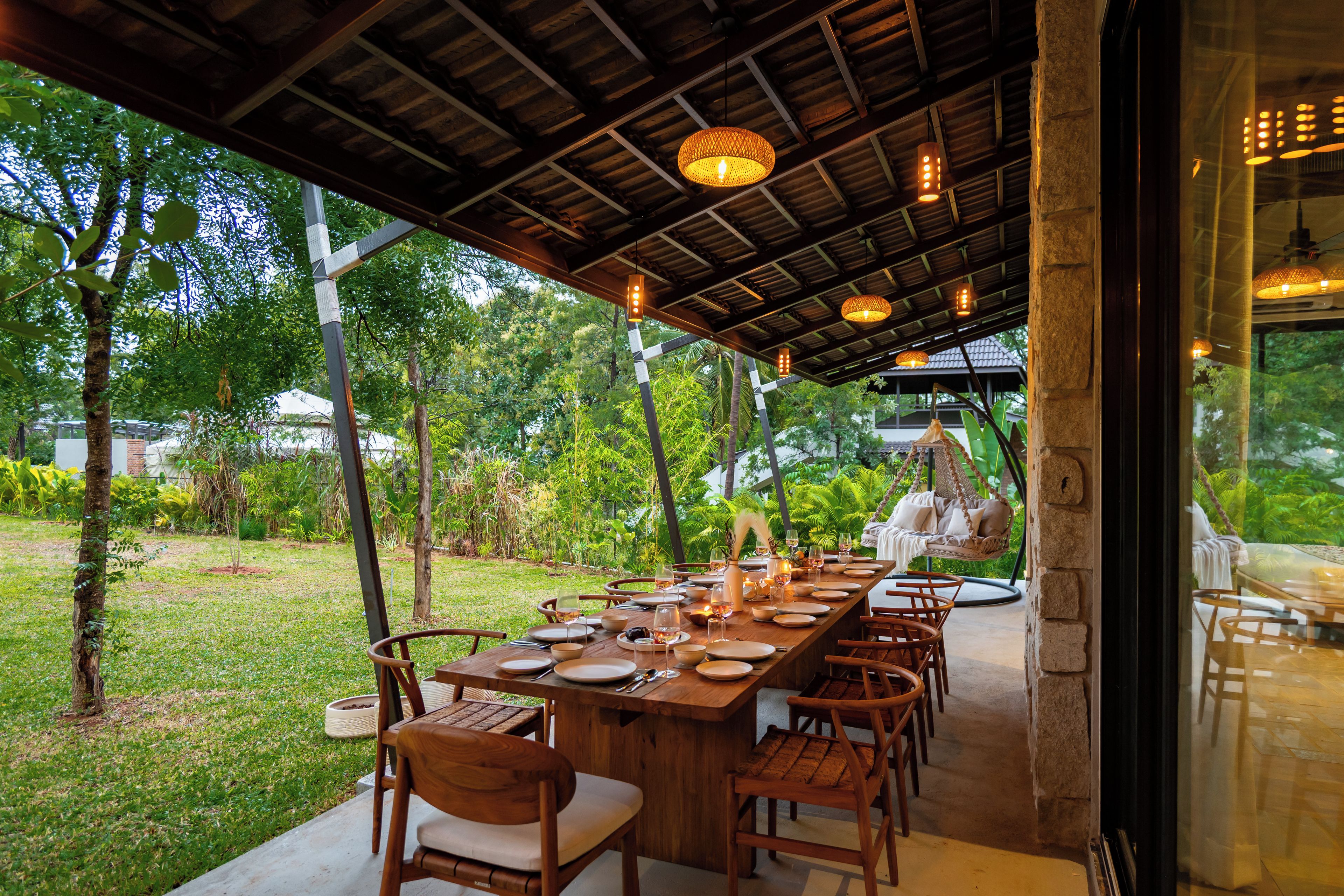 An outdoor dining area with a long wooden table set for a meal, surrounded by chairs, under a covered roof with hanging lights.