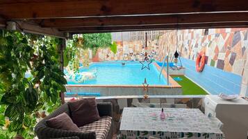 A pool area with a view of a cityscape, a wicker sofa, a table with a patterned tablecloth, and a tiled wall.