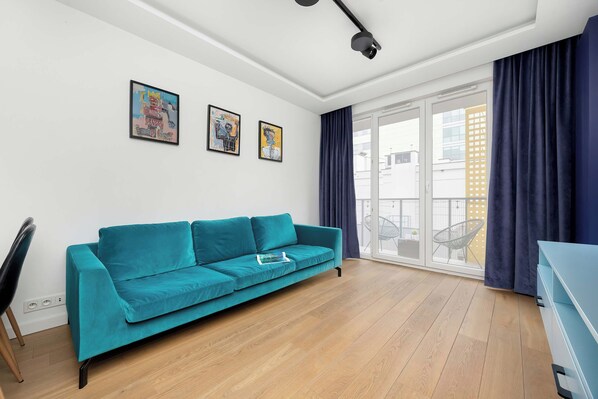 Ein modernes Wohnzimmer mit einem türkisfarbenen Sofa, einem Esstisch und einem großen Fenster mit Vorhängen.