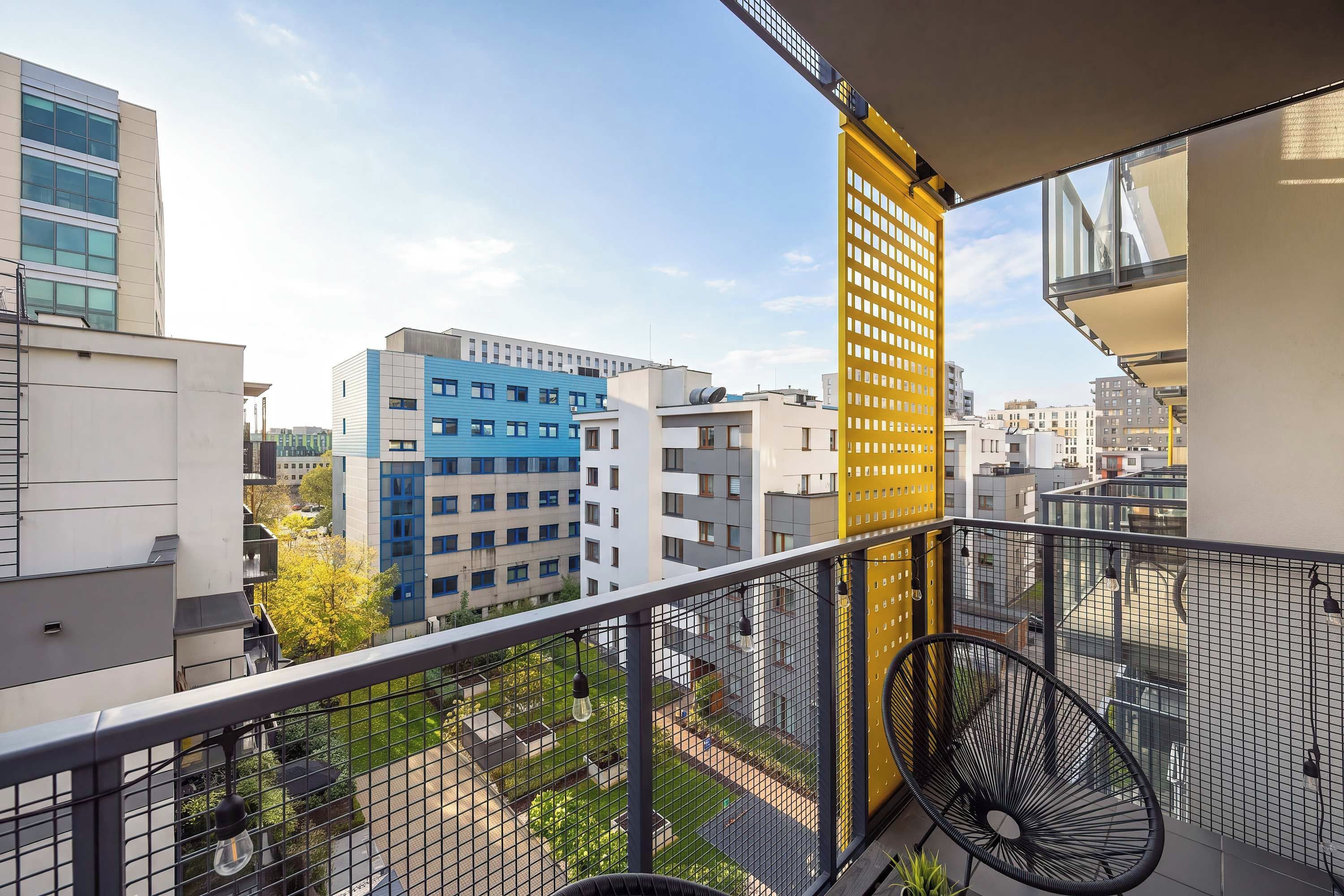 Ein Balkon mit Blick auf umliegende Gebäude, einer gelben, vertikalen Struktur und Sitzgelegenheiten im Freien.