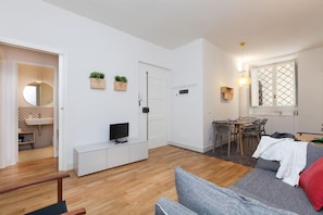 Un espacio habitable moderno con un televisor de pantalla plana sobre un mueble blanco, una zona de comedor con mesa y sillas, y un sofá con un cojín rojo.