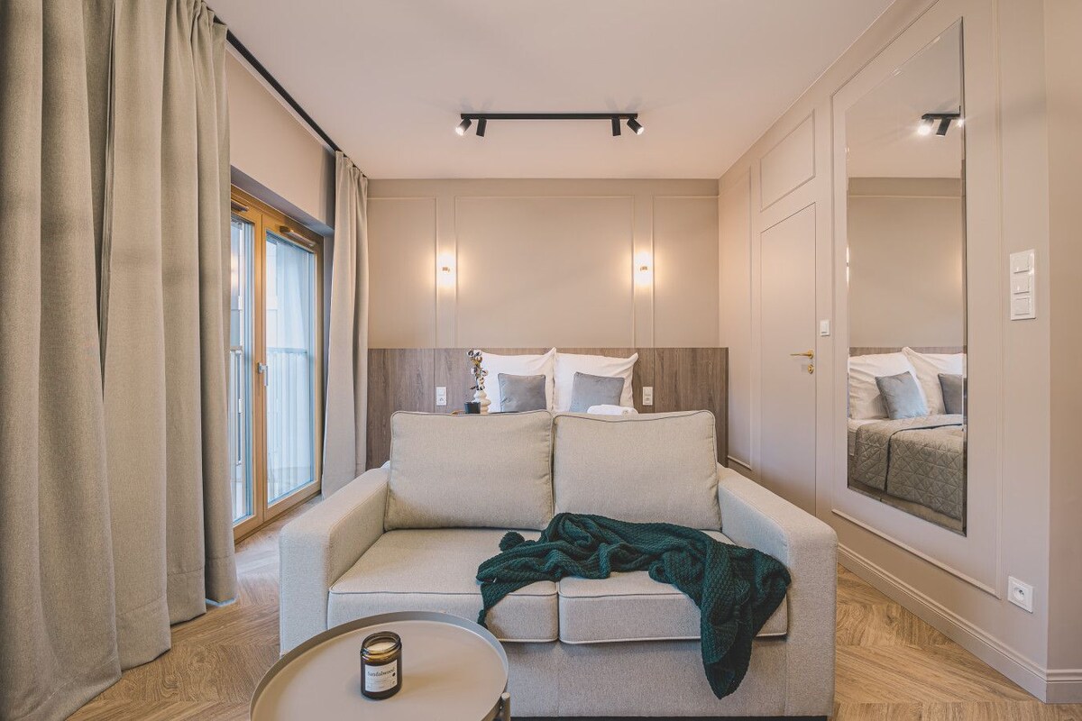 Habitació d'hotel moderna amb un sofà, una taula rodona petita amb una espelma i un mirall gran que reflecteix el llit.