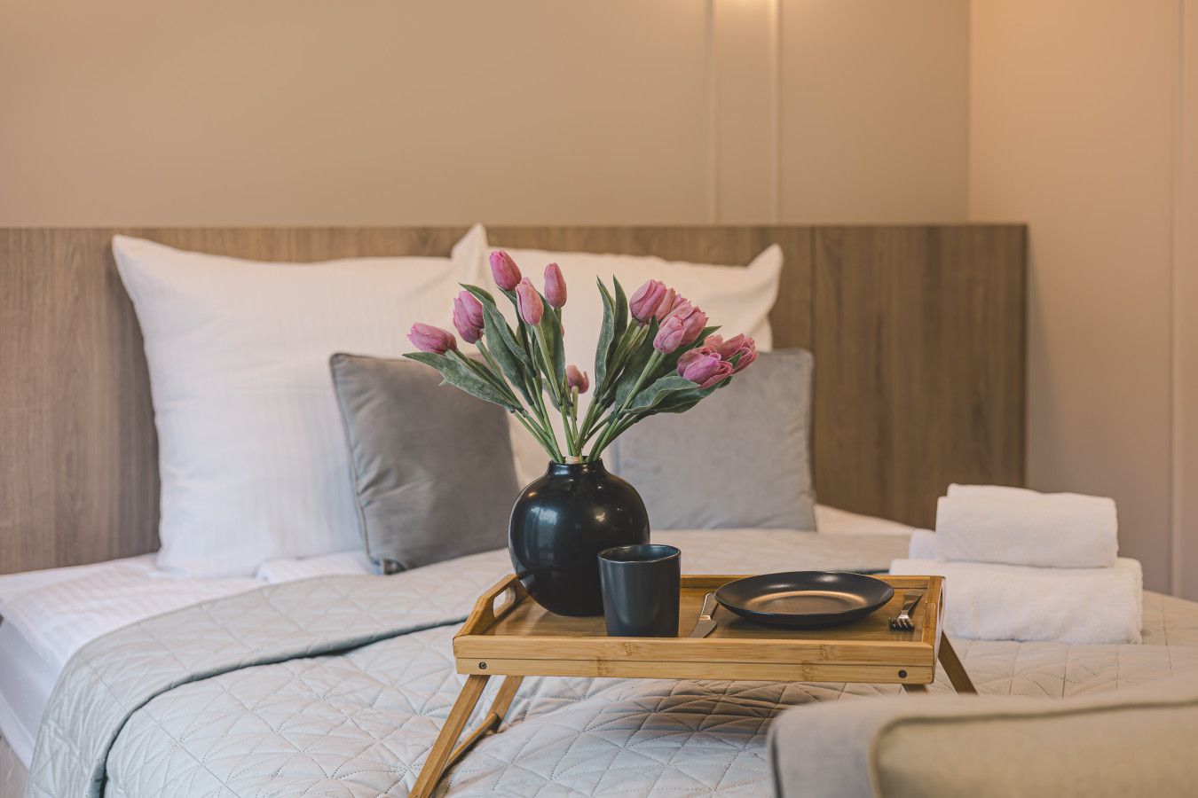Ein Bett mit gesteppter Tagesdecke, ein Nachttisch mit Blumen in einer Vase und ein Tablett mit Teller und Besteck.