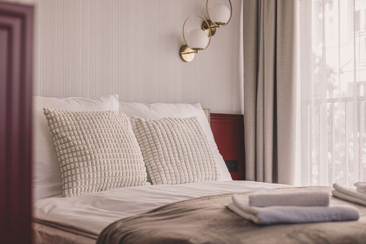 Una habitació d'hotel amb un llit, dos coixins amb textura, una làmpada de nit i una finestra amb cortines.