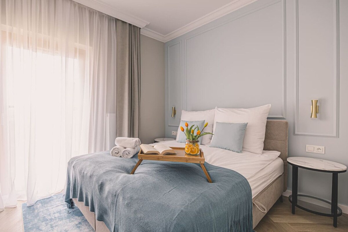 Habitació d'hotel amb un llit, una taula de nit, una taula rodona petita, una finestra amb cortines i un cadi amb tovalloles i un ram de flors.