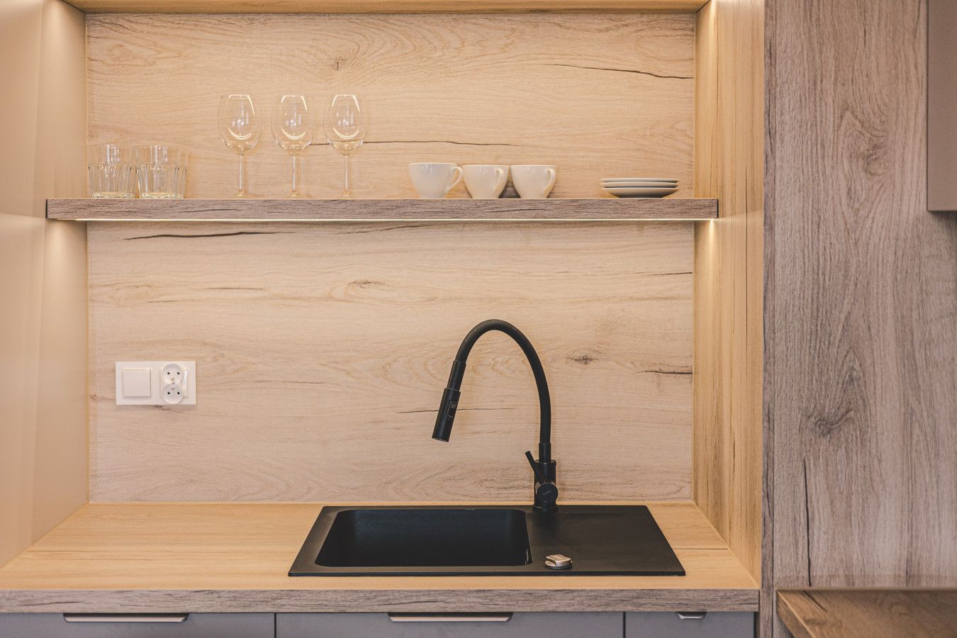 Een moderne keuken met een zwarte spoelbak, een houten aanrechtblad en een plank met glazen en kopjes.