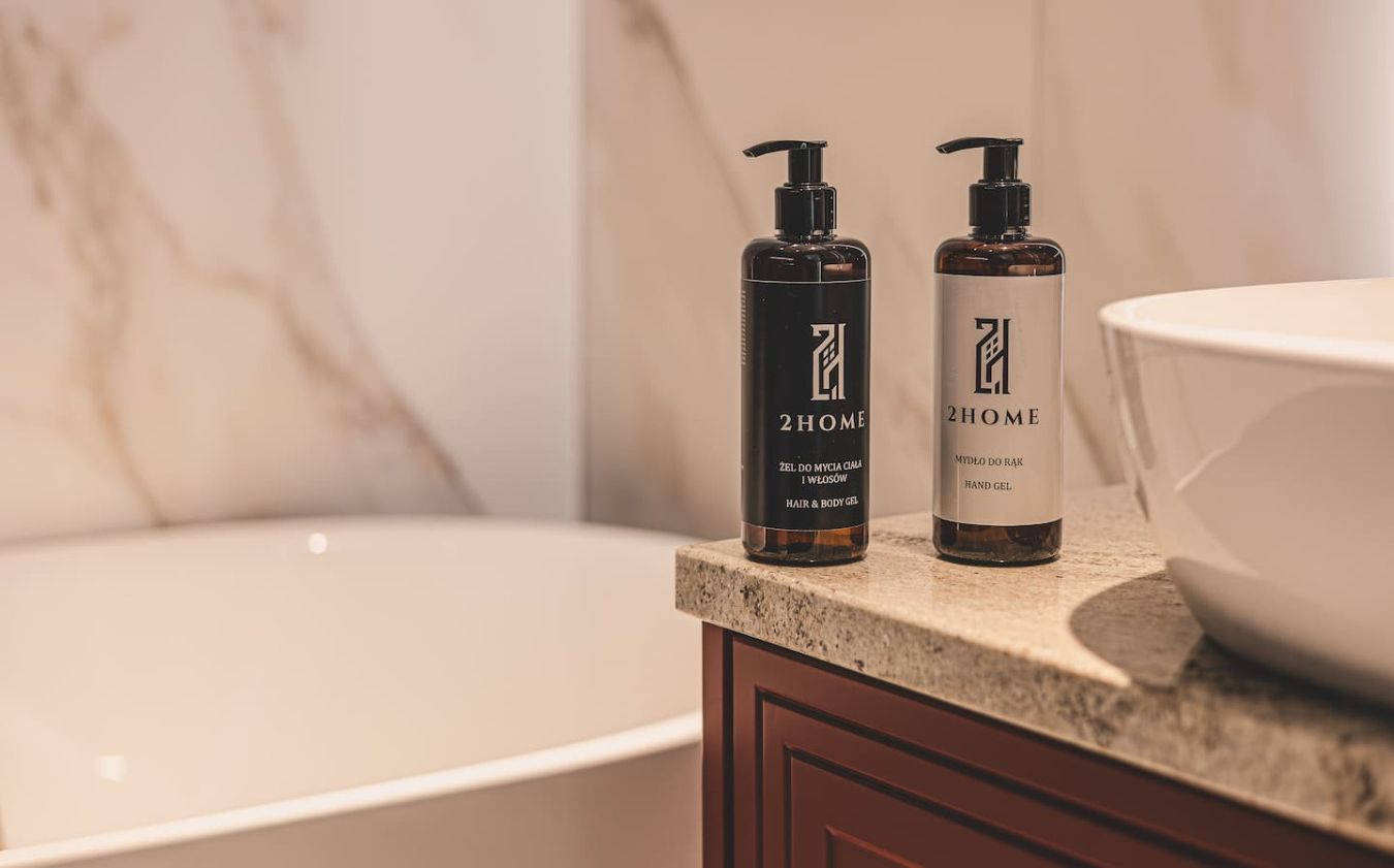 Two bottles of 2HOME hair and body gel on a bathroom countertop next to a bathtub.