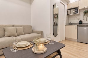 Ein modernes Wohnzimmer mit einer Couch, einem Esstisch gedeckt mit Tellern, Gläsern und einer Kerze, sowie einer Küchenzeile mit Schränken und Geräten.