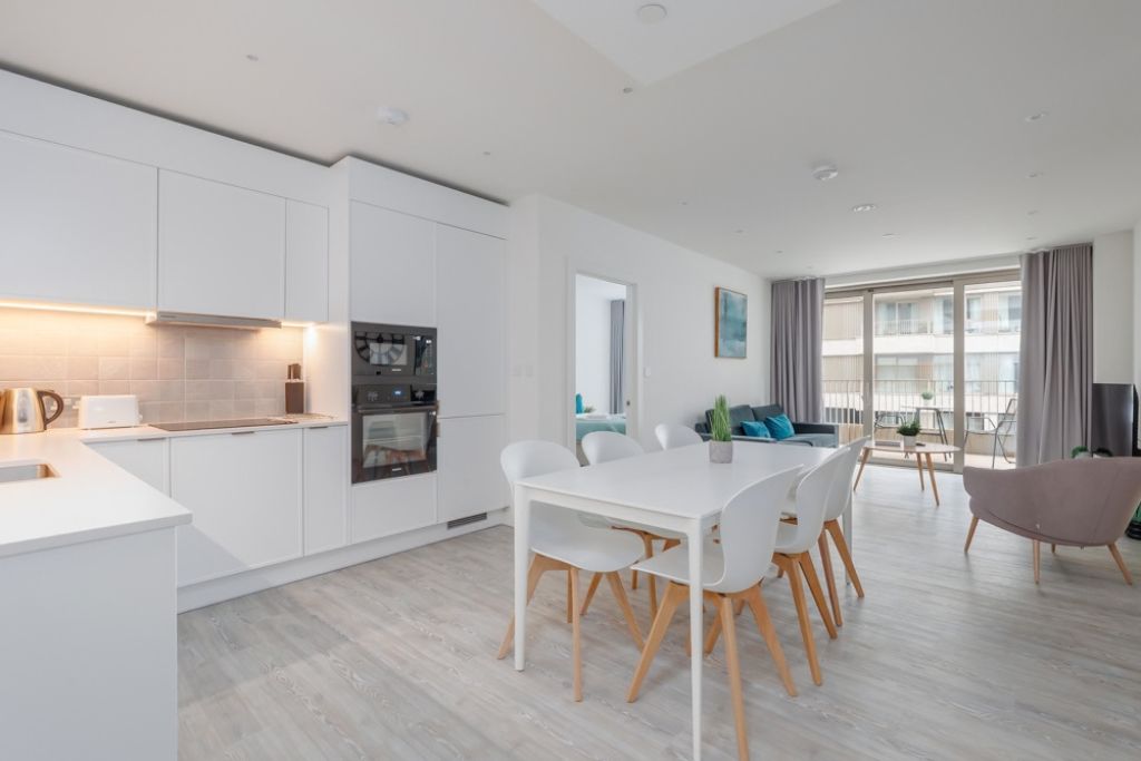 A modern kitchen with white cabinetry, a dining area with a white table and chairs, and a living space with a sofa and a large window.