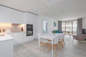 Una cocina moderna con armarios blancos, un comedor con una mesa y sillas blancas, y una sala de estar con un sofá y un ventanal grande.