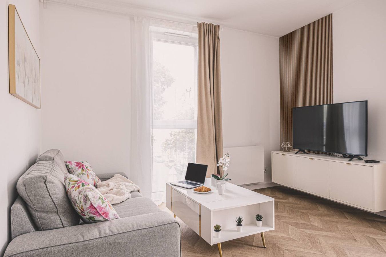 Ein modernes Wohnzimmer mit einem grauen Sofa, einem weißen Couchtisch mit einem Laptop, einem Flachbildfernseher und einem großen Fenster mit Vorhängen.