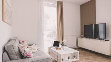 Ein modernes Wohnzimmer mit einem grauen Sofa, einem weißen Couchtisch mit einem Laptop, einem Flachbildfernseher und einem großen Fenster mit Vorhängen.