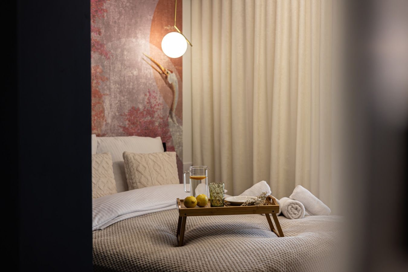 Een opgemaakt bed met een dienblad met citroenen en een glas water, een hangende lamp en een structuurmuur.