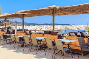 Zona d'estar a l'exterior amb mobles de vimet i paraigües de palla, amb vista a una platja rocosa.