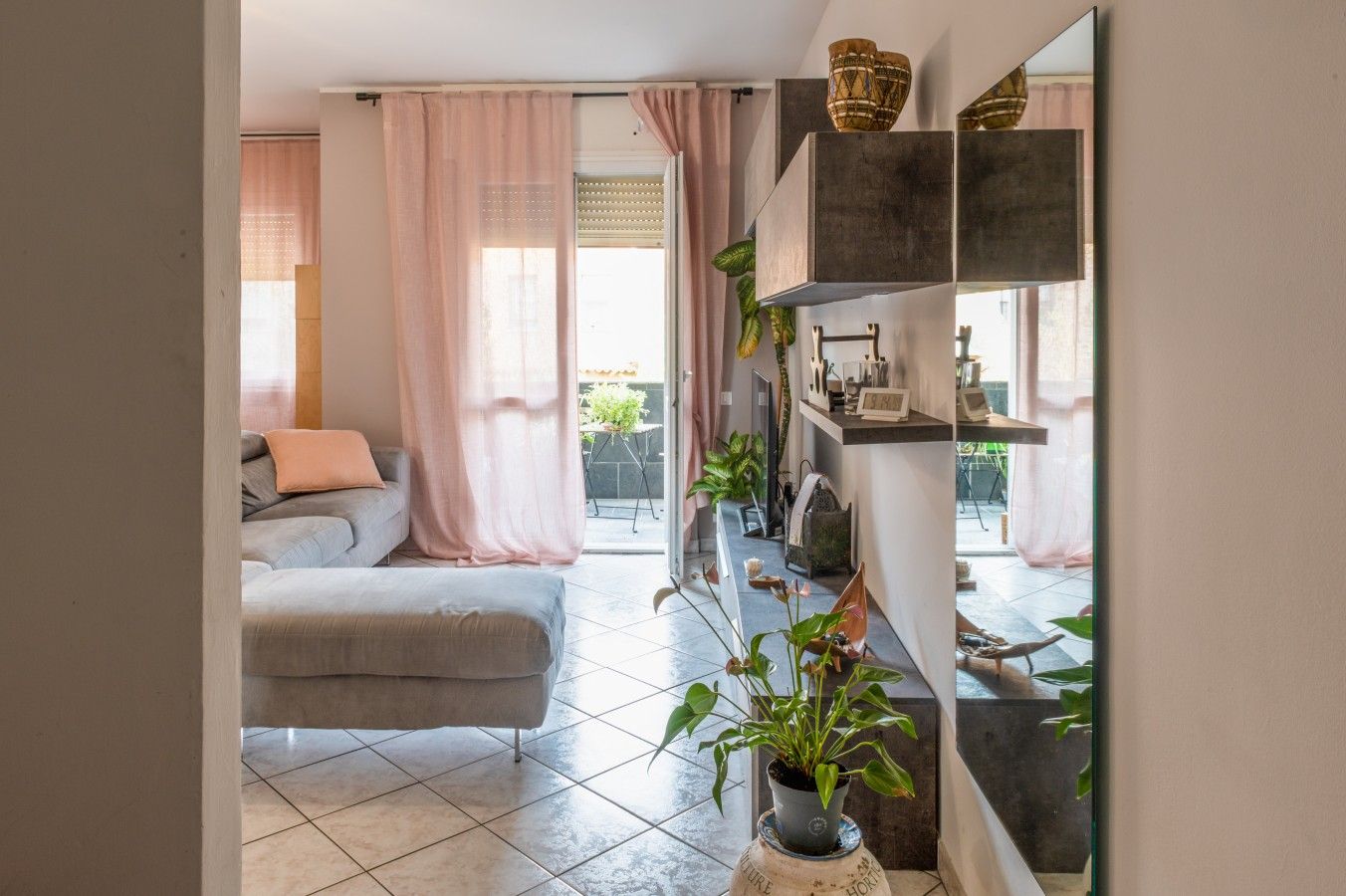 Un salon moderne avec un canapé gris, une table basse et une plante en pot. Une porte-fenêtre coulissante donne accès à un espace extérieur aménagé avec une table et des chaises.