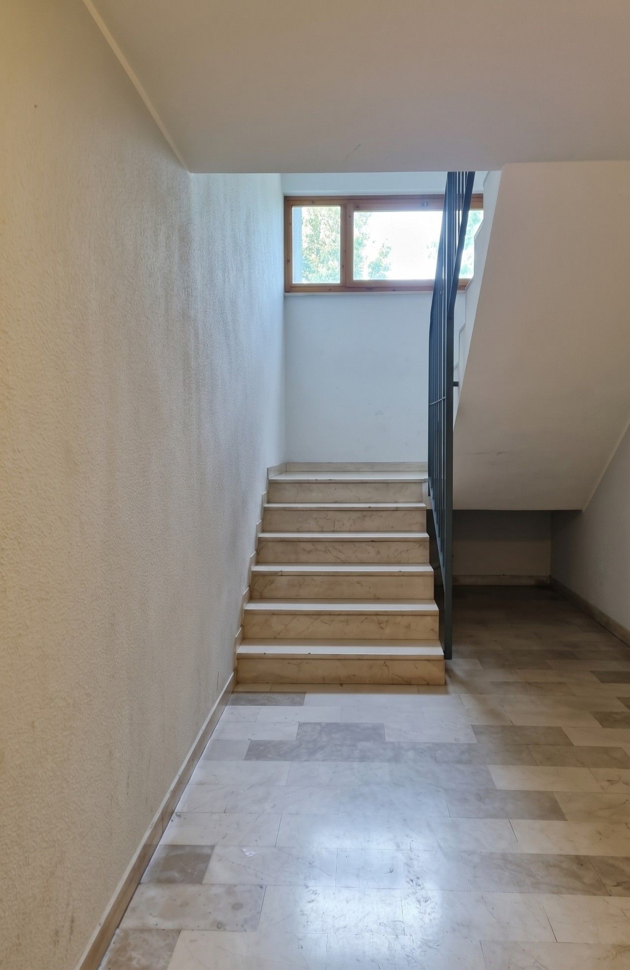 Una escalera con peldaños de madera que conduce a una plataforma, rodeada de paredes lisas y un ventanal en la parte superior.