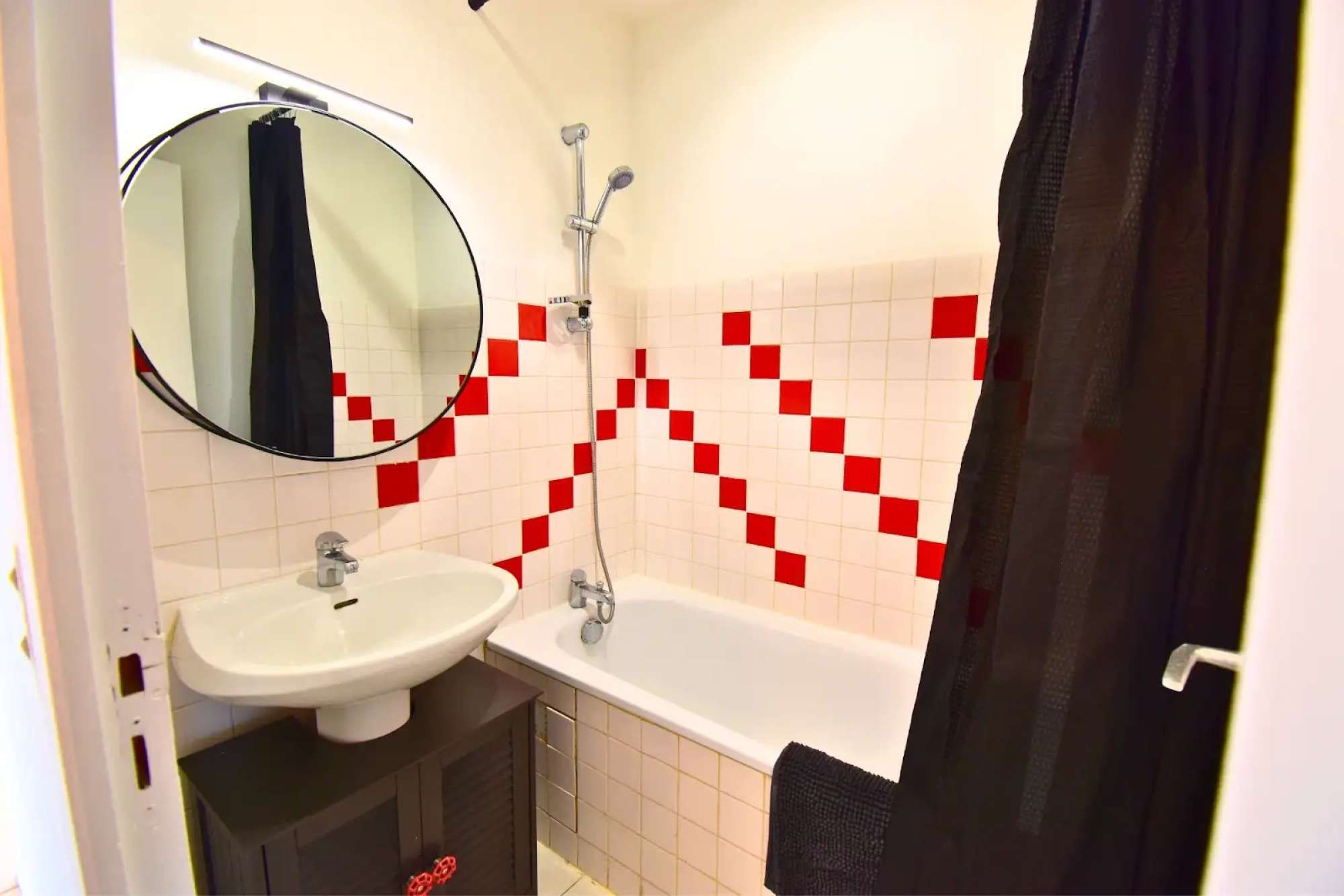 Een badkamer met een wastafel, spiegel, douche en een betegelde muur met een rood-wit patroon.