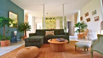Ein Wohnzimmer mit einer Couch, einem runden Couchtisch und einem Korbstuhl. An den Wänden befinden sich Kübelpflanzen und ein Spiegel.