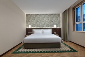 Ein Hotelzimmer mit einem großen Bett, zwei Nachttischlampen, einem Kopfende mit gemustertem Wandpapier, einem Teppich mit grünem Muster und einem Vorhangfenster.
