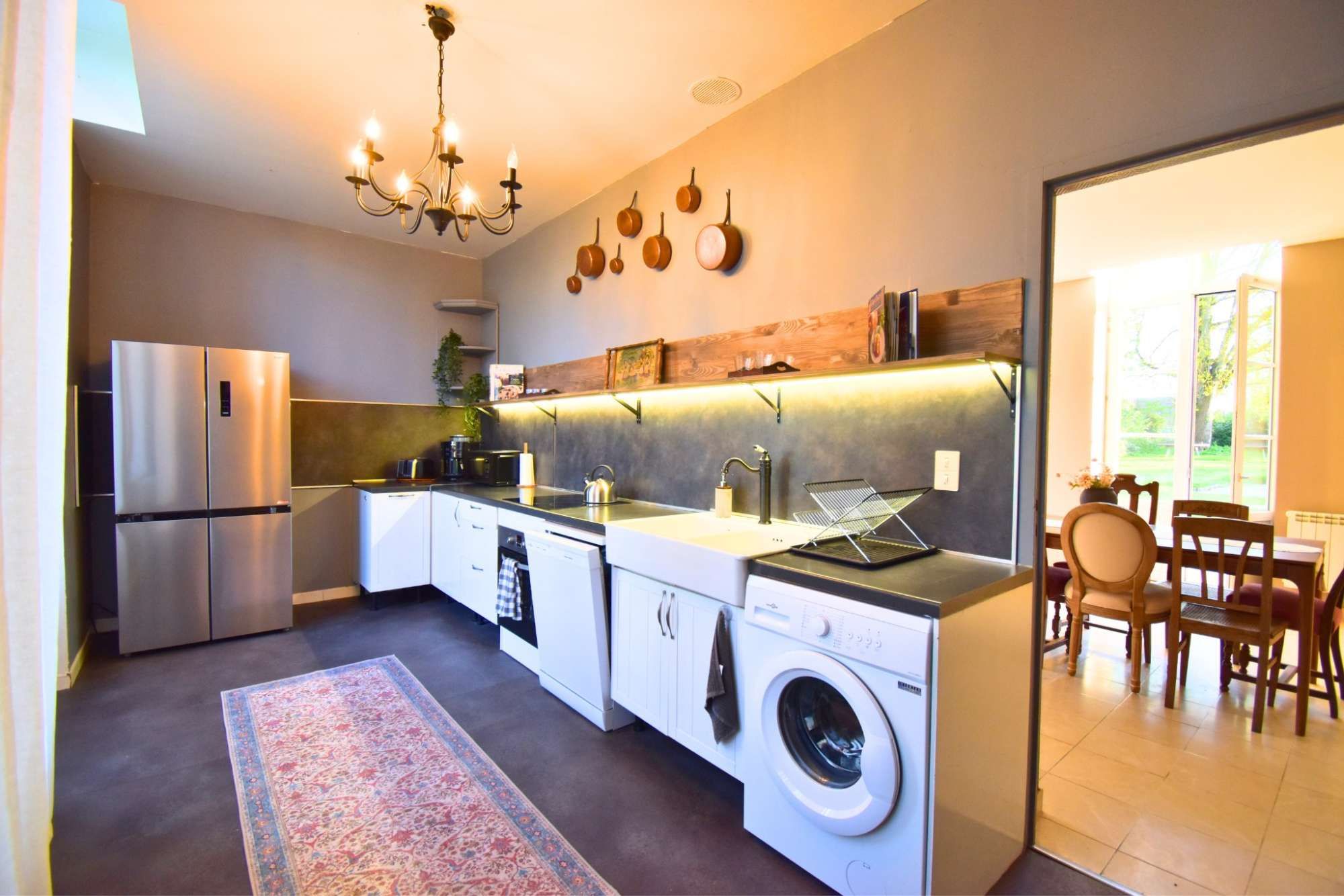 Een moderne keuken met een ingebouwde wasmachine, een kroonluchter en een zichtbare eethoek door een open deur.