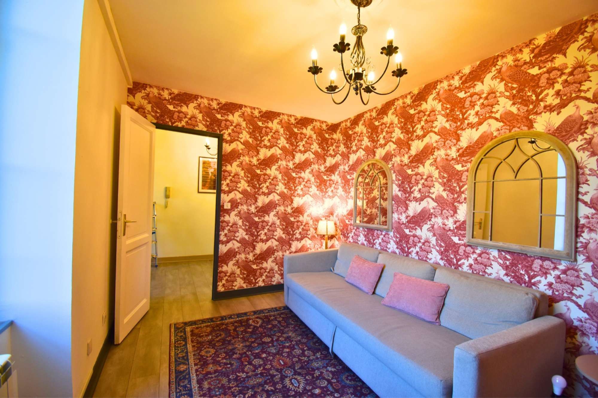 Une pièce avec un papier peint à motifs, un lustre, un canapé avec des coussins, un tapis et un miroir au mur.