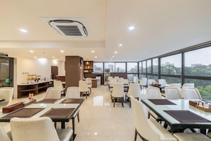A spacious dining area with tables and chairs, a kitchen in the background, and large windows offering a city view. - A25 Hotel 55 Cach Mang Thang 8 (Ho Chi Minh Municipality)