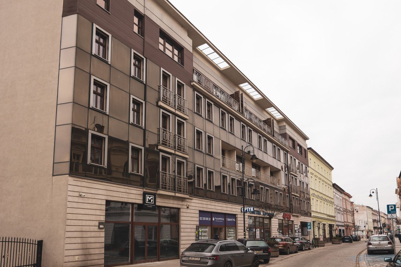 A multi-story building with balconies, a ground-floor entrance, and parked cars along the street.