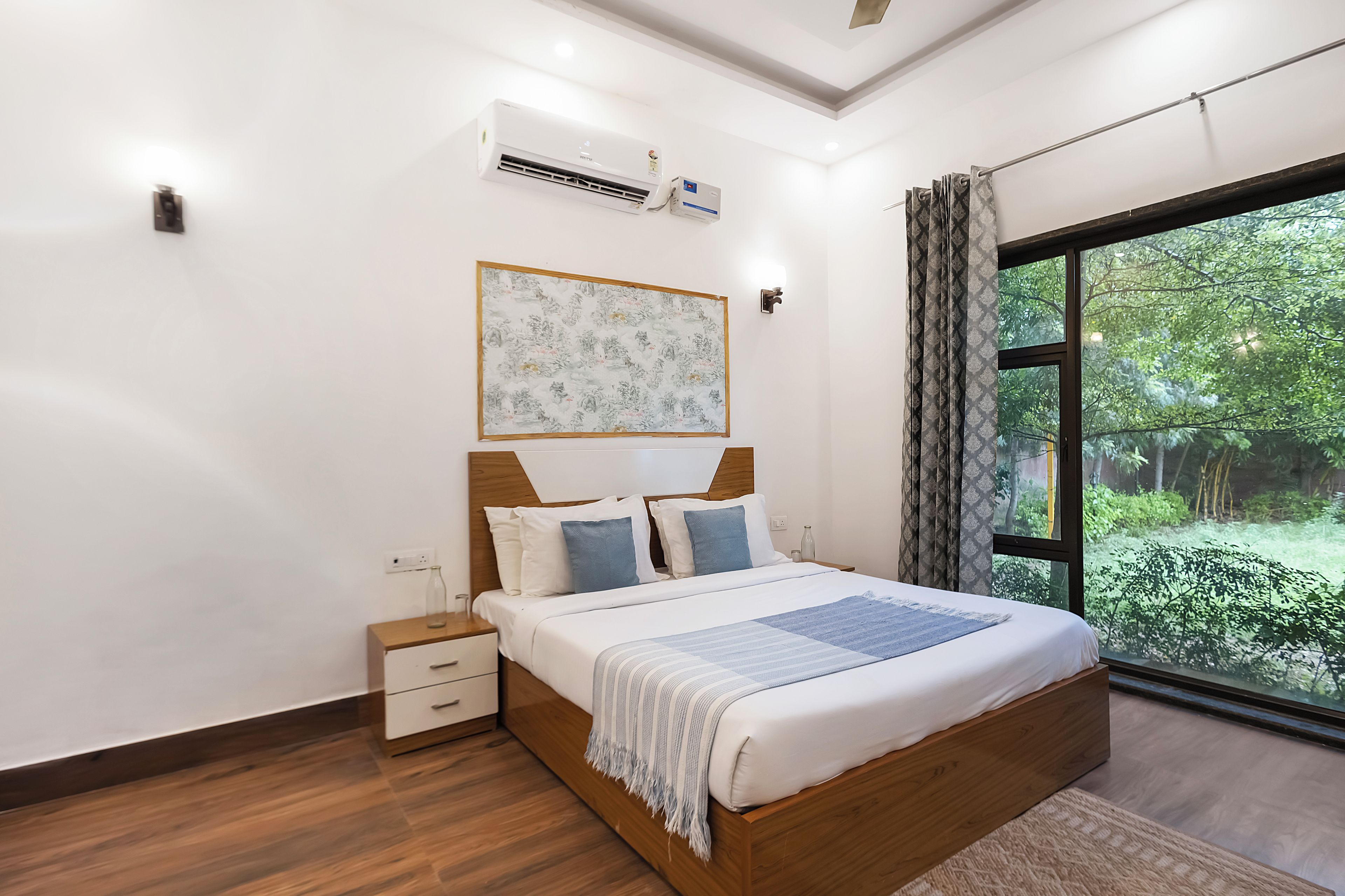 A bedroom with a large bed, a bedside table, a nightstand, a wall-mounted air conditioner, a framed artwork, and a window with curtains.