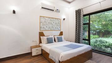A bedroom with a large bed, a bedside table, a nightstand, a wall-mounted air conditioner, a framed artwork, and a window with curtains.