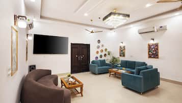 A modern living room with a flat-screen TV, a brown armchair, two blue sofas, a wooden coffee table, and a chandelier.