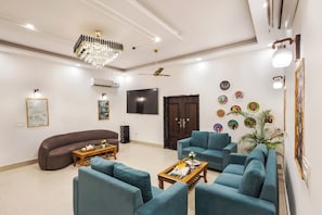 A living room with a chandelier, a flat-screen TV, a sofa, a coffee table, and a wall with decorative plates. - Alaya Stays Aroma Green 4 BHK Farmhouse (Manesar)