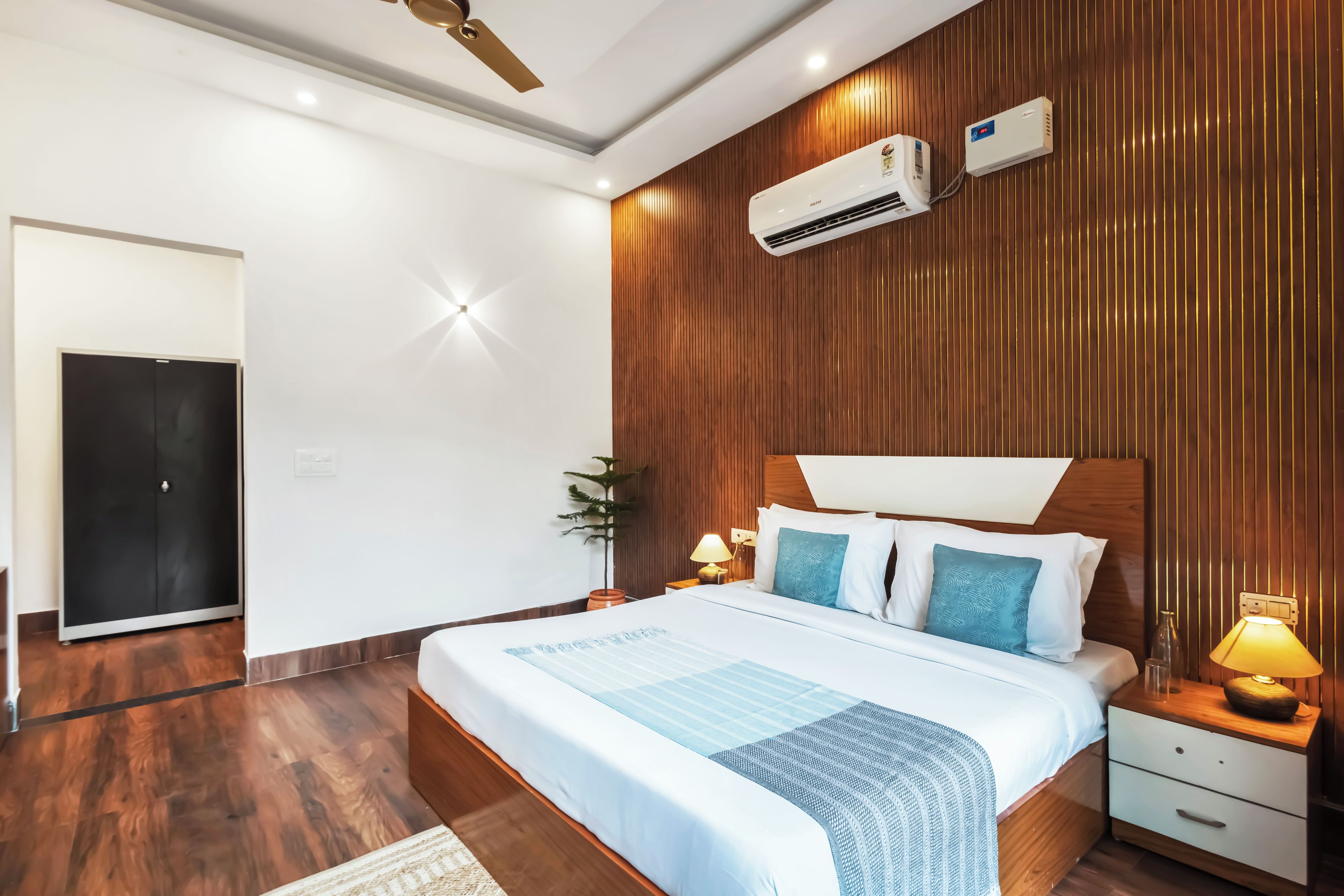 Une chambre moderne avec un mur recouvert de panneaux en bois, un lit doté d’une tête de lit blanche, deux tables de chevet avec des lampes et un appareil de climatisation.