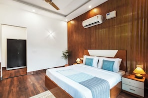 A modern bedroom with a wooden panel wall, a bed with a white headboard, two bedside tables with lamps, and an air conditioning unit. - Alaya Stays Aroma Green 4 BHK Farmhouse (Manesar)