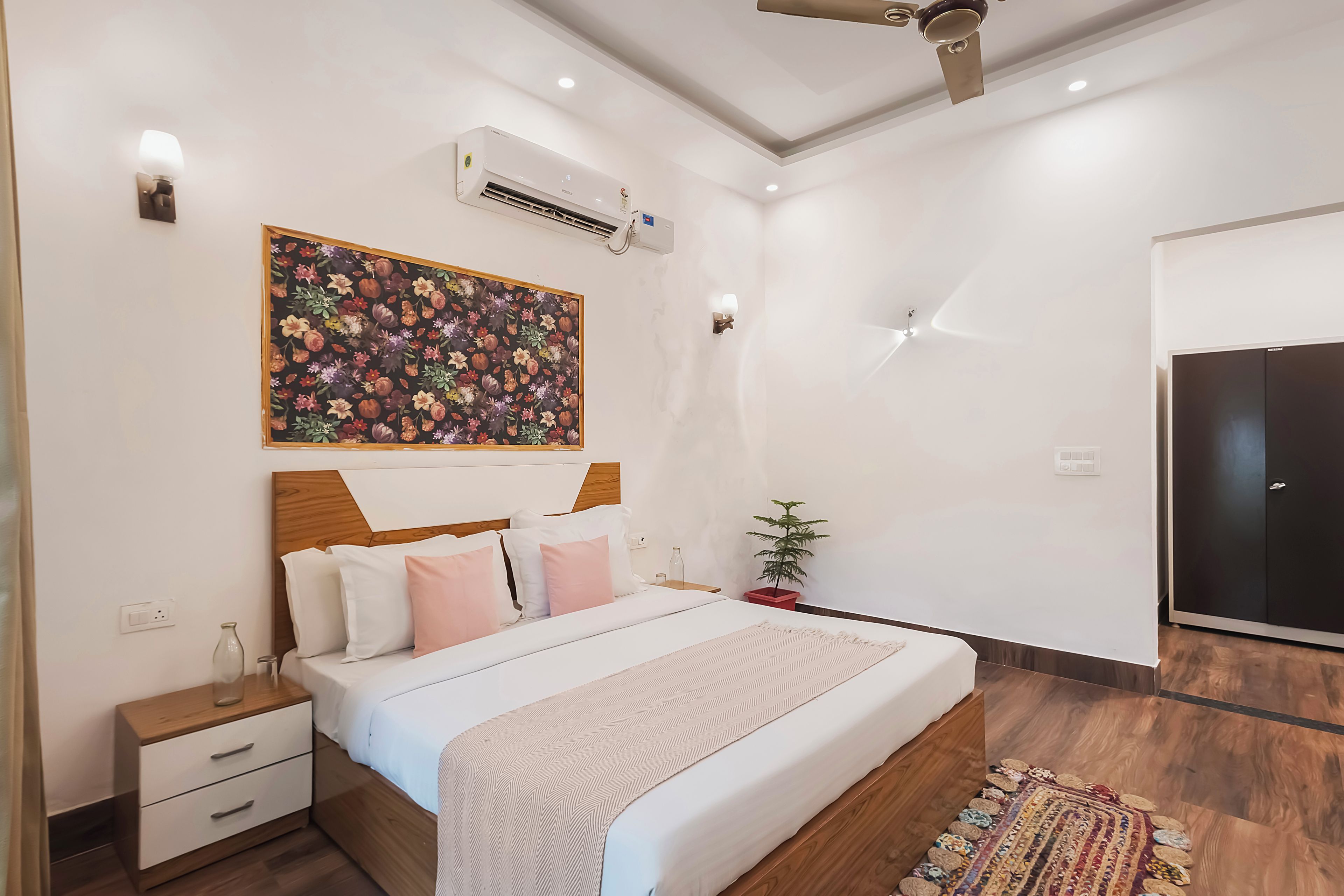 A modern bedroom with a wooden headboard, a large floral artwork, a bed with white and pink bedding, a nightstand, a bedside table, a potted plant, and a wardrobe.