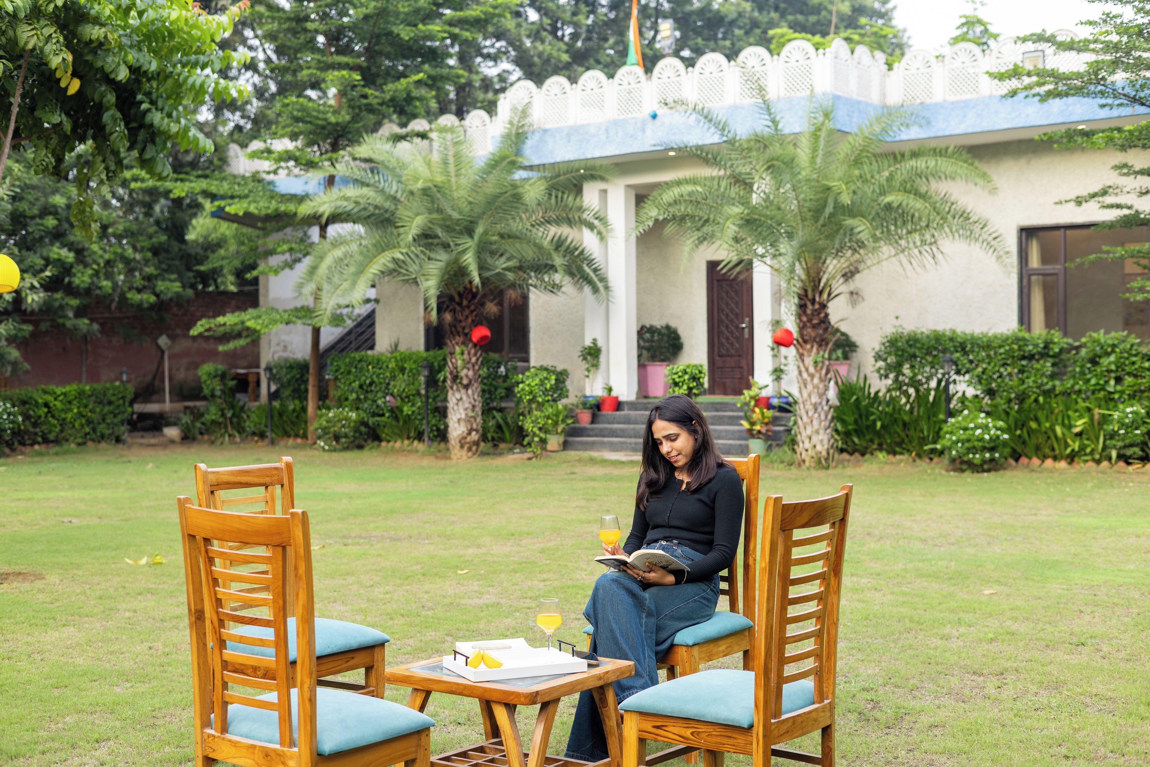 A woman sitting on a wooden bench in a garden with a table, reading a book and holding a glass of orange juice.