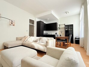 A modern living room with a sectional sofa, a dining area with a wooden table and chairs, and a kitchen with black cabinets and a microwave. - Modern in the Heart of Brussels (Bruxelles)