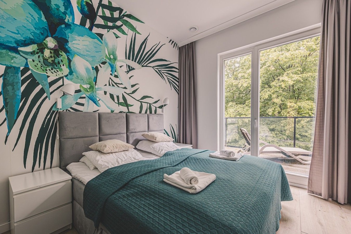 A modern bedroom with a large bed, a nightstand, a wall mural, and a balcony with a view of trees.