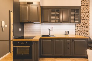 A modern kitchen with dark cabinetry, a stainless steel range hood, a built-in oven, a sink with a faucet, and a stack of firewood against the wall. - Puszcza Białowieska Apartments by 2HomeRent (Nieznany Bór)