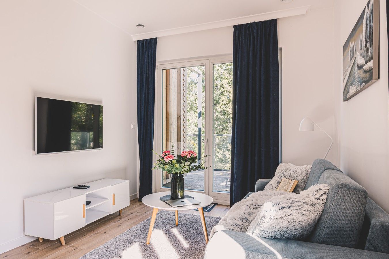 Ein modernes Wohnzimmer mit einem Flachbildfernseher, einer weißen Ablage, einem runden Couchtisch mit Blumenvase, einem grauen Sofa mit weichem Überwurf und einem großen Fenster mit Vorhängen.