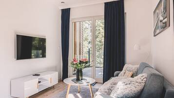 Ein modernes Wohnzimmer mit einem Flachbildfernseher, einer weißen Ablage, einem runden Couchtisch mit Blumenvase, einem grauen Sofa mit weichem Überwurf und einem großen Fenster mit Vorhängen.